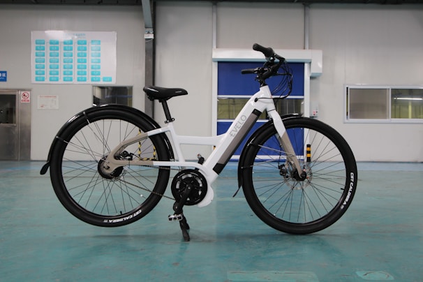 A white electric bicycle stands on a turquoise floor inside a spacious, industrial-like room. The bike features a sleek frame with black tires and a mounted battery. In the background are doors and windows set into light-colored walls, along with a chart or sign on one wall.