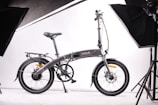 A sleek, modern folding bicycle is positioned against a plain white background, surrounded by studio lighting equipment. The bicycle features a streamlined design, with a dark frame and thin tires, indicating it is built for efficiency and portability.