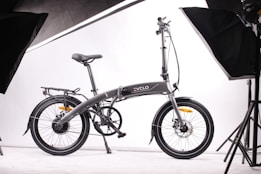 A sleek, modern folding bicycle is positioned against a plain white background, surrounded by studio lighting equipment. The bicycle features a streamlined design, with a dark frame and thin tires, indicating it is built for efficiency and portability.