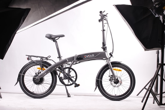 A sleek, modern folding bicycle is positioned against a plain white background, surrounded by studio lighting equipment. The bicycle features a streamlined design, with a dark frame and thin tires, indicating it is built for efficiency and portability.