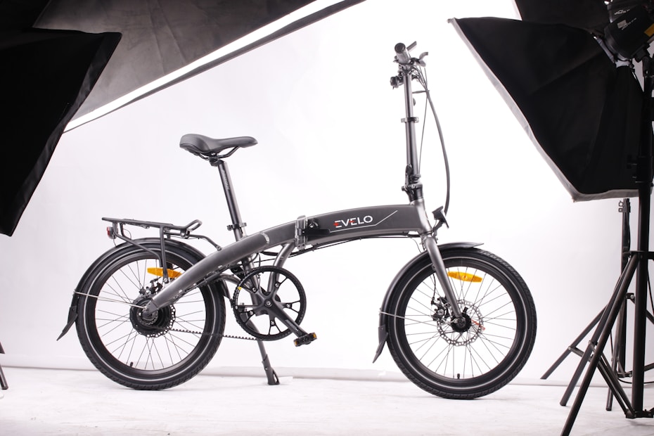 A sleek, modern folding bicycle is positioned against a plain white background, surrounded by studio lighting equipment. The bicycle features a streamlined design, with a dark frame and thin tires, indicating it is built for efficiency and portability.