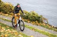 A person is happily riding a bicycle along a path surrounded by lush greenery and fallen autumn leaves. The background features an expansive view of a lake or river, adding a serene natural setting.