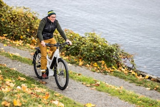 A person is happily riding a bicycle along a path surrounded by lush greenery and fallen autumn leaves. The background features an expansive view of a lake or river, adding a serene natural setting.