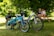 Two EVELO branded electric bikes are parked on a grassy area under the shade of trees. A couple is sitting on a picnic bench in the background, creating a relaxed and peaceful atmosphere. The scene suggests a leisurely outdoor setting, ideal for biking and enjoying nature.