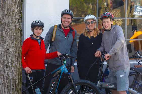A group of friends on cruiser bikes, smiling and waving near a rustic coffee shop with dogs nearby.