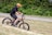A person wearing a bright orange helmet rides a black and orange electric bicycle on a grassy path. The background includes lush green foliage and a distant gravel pathway. The individual appears to be enjoying an outdoor activity.