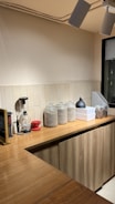 A cozy kitchen counter with wooden cabinets and a tiled backsplash. Glass jars filled with grains or coffee beans are neatly arranged on the counter alongside a coffee grinder and a red coffee dripper. Industrial-style ceiling lights provide illumination to the area.