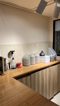 A cozy kitchen counter with wooden cabinets and a tiled backsplash. Glass jars filled with grains or coffee beans are neatly arranged on the counter alongside a coffee grinder and a red coffee dripper. Industrial-style ceiling lights provide illumination to the area.