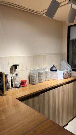 A cozy kitchen counter with wooden cabinets and a tiled backsplash. Glass jars filled with grains or coffee beans are neatly arranged on the counter alongside a coffee grinder and a red coffee dripper. Industrial-style ceiling lights provide illumination to the area.