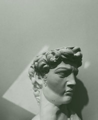 A stone bust of a classical male figure with curly hair, captured in a monochromatic green tone. The sculpture is shown from a side angle, highlighting the intricate details of the hair and facial features. The lighting creates shadows that enhance the three-dimensional aspect of the artwork.