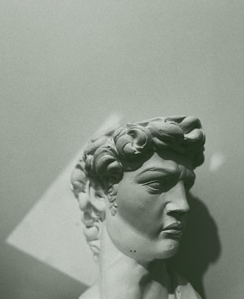 A stone bust of a classical male figure with curly hair, captured in a monochromatic green tone. The sculpture is shown from a side angle, highlighting the intricate details of the hair and facial features. The lighting creates shadows that enhance the three-dimensional aspect of the artwork.