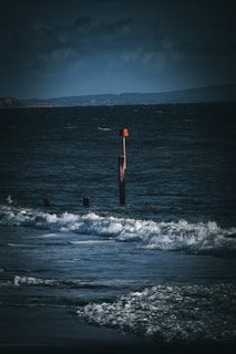 Dark and moody ocean scene with waves crashing onto the shore. A wooden groyne with an orange navigation marker stands prominently in the water, while dark clouds loom in the background.