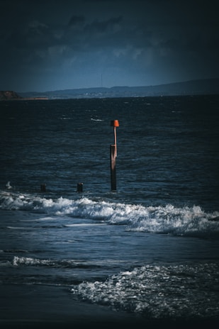 Dark and moody ocean scene with waves crashing onto the shore. A wooden groyne with an orange navigation marker stands prominently in the water, while dark clouds loom in the background.