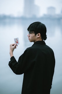 A person with short black hair and glasses is holding a playing card with an illustrated figure in a red and blue outfit. The background features a serene body of water, creating a calm atmosphere.
