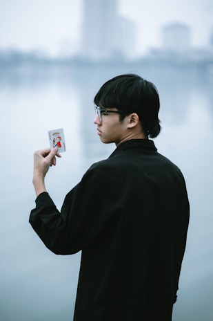 A person with short black hair and glasses is holding a playing card with an illustrated figure in a red and blue outfit. The background features a serene body of water, creating a calm atmosphere.