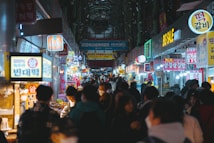 A bustling market scene with vibrant signs and lights hanging above numerous food stalls. People are walking and browsing along both sides of a narrow street. The atmosphere appears lively and crowded, capturing interactions among visitors and vendors.