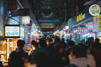 A bustling market scene with vibrant signs and lights hanging above numerous food stalls. People are walking and browsing along both sides of a narrow street. The atmosphere appears lively and crowded, capturing interactions among visitors and vendors.