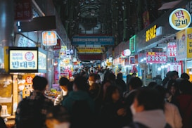 A bustling market scene with vibrant signs and lights hanging above numerous food stalls. People are walking and browsing along both sides of a narrow street. The atmosphere appears lively and crowded, capturing interactions among visitors and vendors.