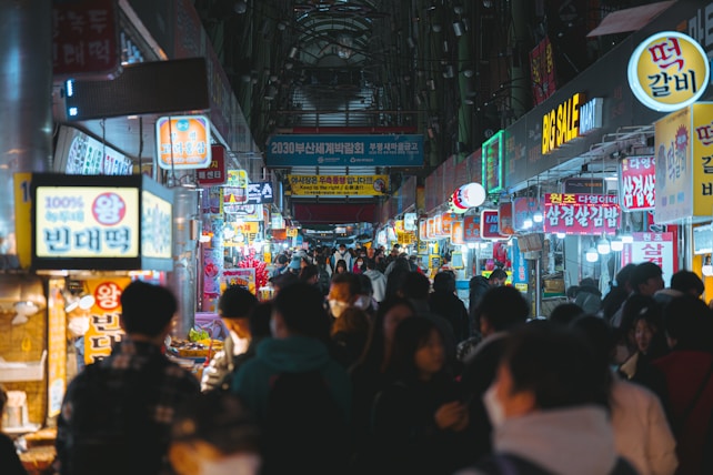 A bustling market scene with vibrant signs and lights hanging above numerous food stalls. People are walking and browsing along both sides of a narrow street. The atmosphere appears lively and crowded, capturing interactions among visitors and vendors.