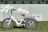 A white electric tricycle with a brown seat is parked on green grass in front of a light blue wall. The tricycle features a large basket at the rear and is equipped with three large wheels. The wall behind is decorated with a mural of white birds and a stylized blue fish. Circular windows are visible along the top of the wall.
