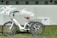 A white electric tricycle with a brown seat is parked on green grass in front of a light blue wall. The tricycle features a large basket at the rear and is equipped with three large wheels. The wall behind is decorated with a mural of white birds and a stylized blue fish. Circular windows are visible along the top of the wall.
