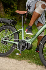 A person with knee pads resting on a green e-bike. The bike features a brown seat and thick tires. The individual is wearing shorts and sneakers that have colorful stripes. In the background, there is grassy terrain and gravel on the path.