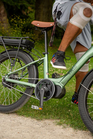 A person with knee pads resting on a green e-bike. The bike features a brown seat and thick tires. The individual is wearing shorts and sneakers that have colorful stripes. In the background, there is grassy terrain and gravel on the path.
