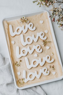 Four cookies in the shape of the word 'love' are neatly arranged on a baking tray with parchment paper. Small, delicate white flowers with pink centers are scattered around the cookies, adding a decorative touch. The cookies are coated with white icing, and additional small heart-shaped cookies accompany them. The background consists of a soft white fabric.