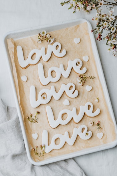Four cookies in the shape of the word 'love' are neatly arranged on a baking tray with parchment paper. Small, delicate white flowers with pink centers are scattered around the cookies, adding a decorative touch. The cookies are coated with white icing, and additional small heart-shaped cookies accompany them. The background consists of a soft white fabric.