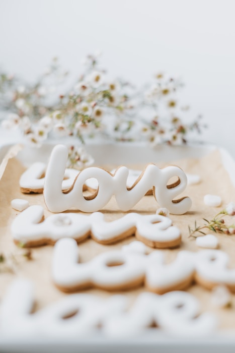 Cookies shaped like the word 'love' are decorated with white icing and arranged on a baking sheet lined with parchment paper. In the background, there are delicate white flowers with small blossoms, creating a soft and romantic atmosphere.