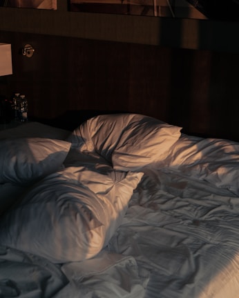 A dimly lit bedroom scene features an unmade bed with its white sheets and pillows tossed carelessly. The bed is notably wrinkled, suggesting recent use or rest, and the lighting casts soft shadows, accentuating the folds of the fabric. Two water bottles sit on a small nightstand beside the bed.