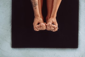 A person is performing a yoga pose, gripping their toes with both hands while sitting on a black yoga mat. Visible tattoos are on one arm, and the focus is on the feet and hands against a textured surface.