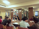 A group of people is seated around a large wooden table in a library, engaging in discussion or study. The background features shelves filled with books, labeled with signs like 'Silence Zone', indicating different sections such as 'B.TECH' and 'BJMC'. The room is well-lit with artificial lighting, and there are windows with blue curtains.