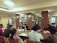 A group of people is seated around a large wooden table in a library, engaging in discussion or study. The background features shelves filled with books, labeled with signs like 'Silence Zone', indicating different sections such as 'B.TECH' and 'BJMC'. The room is well-lit with artificial lighting, and there are windows with blue curtains.