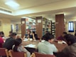 A group of people is seated around a large wooden table in a library, engaging in discussion or study. The background features shelves filled with books, labeled with signs like 'Silence Zone', indicating different sections such as 'B.TECH' and 'BJMC'. The room is well-lit with artificial lighting, and there are windows with blue curtains.