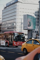 A red bus is on the road in an urban area, with a yellow taxi nearby. There's a large glass office building in the background, and an electronic billboard displaying an advertisement featuring people in medical attire. Street signs direct traffic to various city districts.