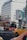 A red bus is on the road in an urban area, with a yellow taxi nearby. There's a large glass office building in the background, and an electronic billboard displaying an advertisement featuring people in medical attire. Street signs direct traffic to various city districts.