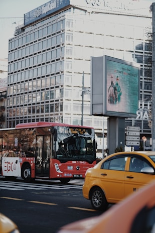 A red bus is on the road in an urban area, with a yellow taxi nearby. There's a large glass office building in the background, and an electronic billboard displaying an advertisement featuring people in medical attire. Street signs direct traffic to various city districts.