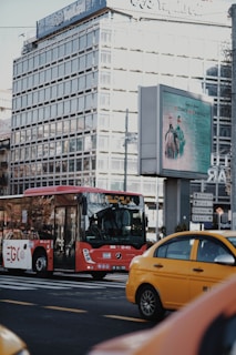 A red bus is on the road in an urban area, with a yellow taxi nearby. There's a large glass office building in the background, and an electronic billboard displaying an advertisement featuring people in medical attire. Street signs direct traffic to various city districts.