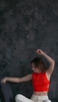 A person wearing a bright red sleeveless top and white pants appears to be moving energetically against a dark, textured backdrop. Their arms are raised, suggesting a dance or expressive motion.