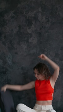 A person wearing a bright red sleeveless top and white pants appears to be moving energetically against a dark, textured backdrop. Their arms are raised, suggesting a dance or expressive motion.
