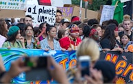 A large group of people participating in a protest or demonstration, holding colorful banners and signs with environmental messages. Some individuals have painted faces and are wearing traditional or cultural attire.