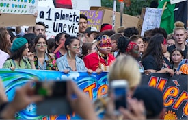 A large group of people participating in a protest or demonstration, holding colorful banners and signs with environmental messages. Some individuals have painted faces and are wearing traditional or cultural attire.