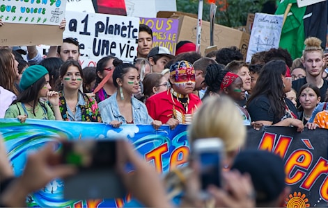 A large group of people participating in a protest or demonstration, holding colorful banners and signs with environmental messages. Some individuals have painted faces and are wearing traditional or cultural attire.
