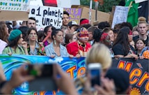 A large group of people participating in a protest or demonstration, holding colorful banners and signs with environmental messages. Some individuals have painted faces and are wearing traditional or cultural attire.