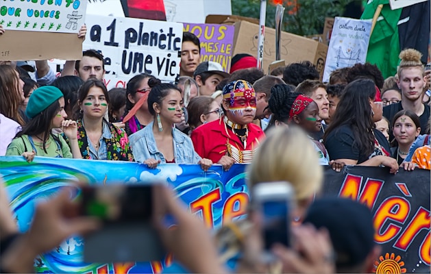 A large group of people participating in a protest or demonstration, holding colorful banners and signs with environmental messages. Some individuals have painted faces and are wearing traditional or cultural attire.