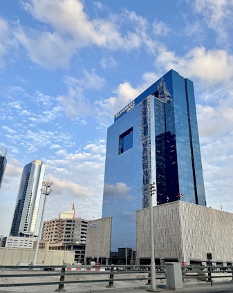A series of modern skyscrapers stand under a blue sky with scattered clouds. The tall glass buildings reflect the sky, with the largest building prominently displaying 'LANDMARK GROUP' on its facade. Surrounding structures vary in height and design, adding to the urban cityscape.