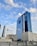 A series of modern skyscrapers stand under a blue sky with scattered clouds. The tall glass buildings reflect the sky, with the largest building prominently displaying 'LANDMARK GROUP' on its facade. Surrounding structures vary in height and design, adding to the urban cityscape.