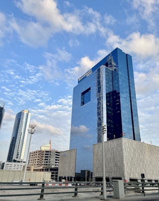 A series of modern skyscrapers stand under a blue sky with scattered clouds. The tall glass buildings reflect the sky, with the largest building prominently displaying 'LANDMARK GROUP' on its facade. Surrounding structures vary in height and design, adding to the urban cityscape.