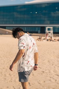 A person wearing a white shirt with abstract patterns and gray shorts walks along a sandy beach. A large modern building with a glass facade is in the background, along with a lifeguard stand and several people lounging on the sand.